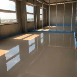 Świeżo wylana, gładka posadzka w jasnym odcieniu w surowym wnętrzu budynku w Kaliszu, zabezpieczona niebieską taśmą przy ścianach i metalowych profilach.