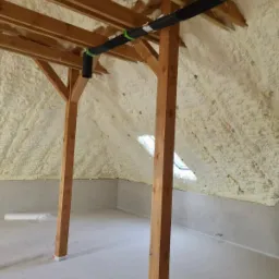 Poddasze po ociepleniu pianką, z widoczną drewnianą konstrukcją dachu i czarną rurą wentylacyjną pod sufitem.