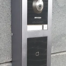 Szary wideodomofon firmy Hikvision zamontowany na szarej, chropowatej ścianie zewnętrznej budynku.