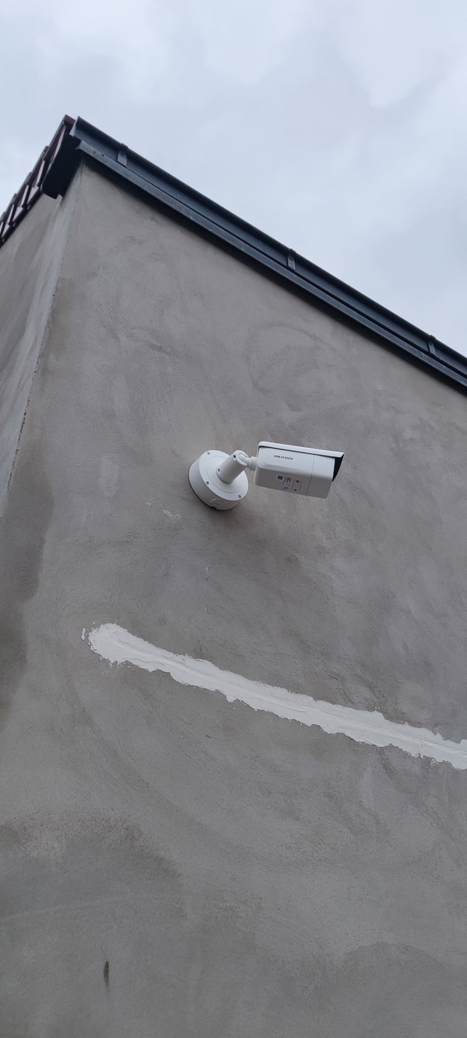Biała kamera monitoringu IP marki Hikvision zamontowana na szarej, surowej ścianie budynku, widoczna fuga wypełniona białą masą uszczelniającą, pochmurne niebo w tle.