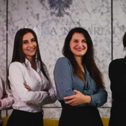 Zespół notarialny: cztery uśmiechnięte kobiety w eleganckich strojach stojące na tle marmurowej ściany z godłem państwowym i napisem 'Izba Notarialna'