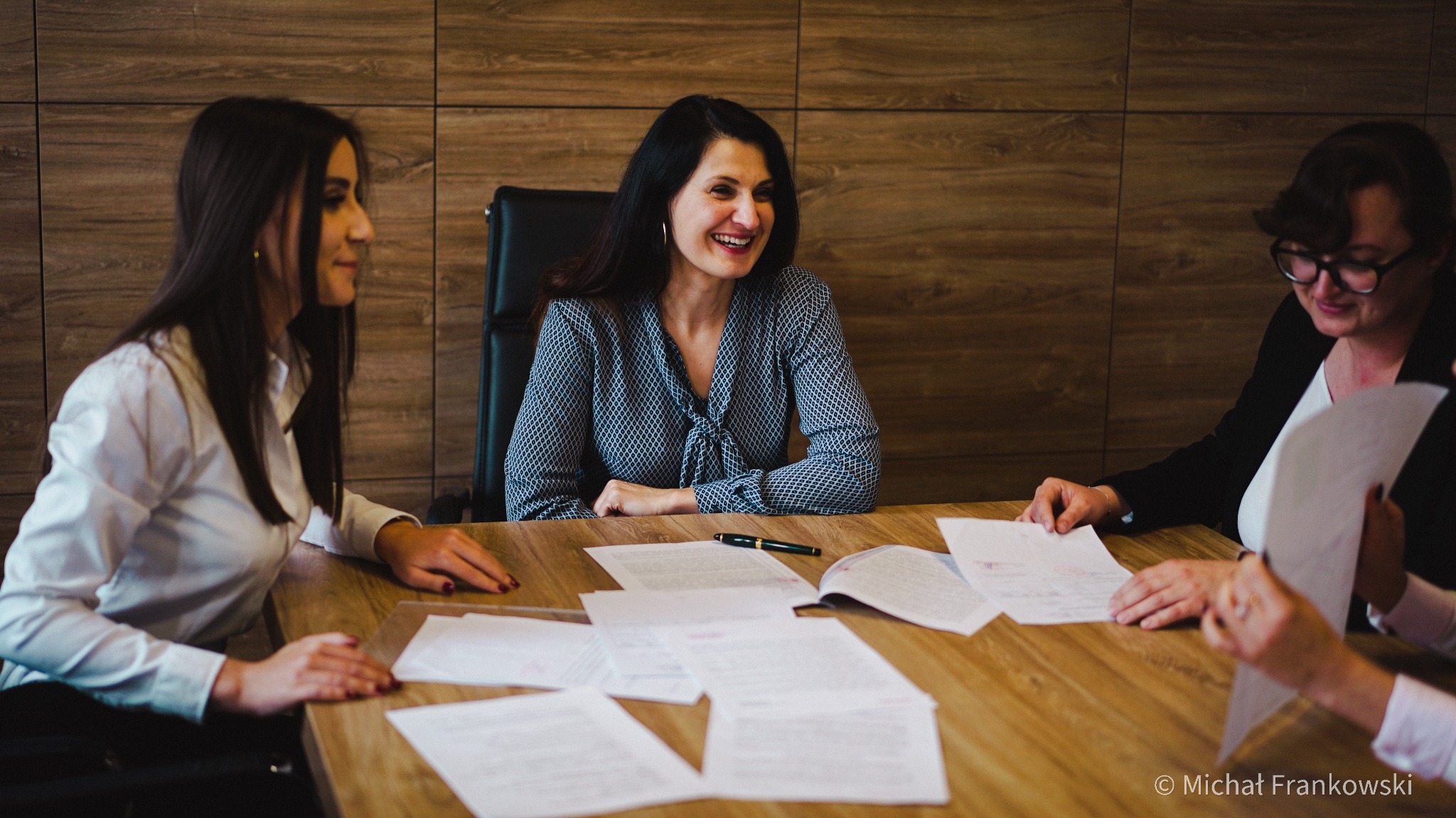 Trzy kobiety przy drewnianym stole analizują dokumenty, na stole leży pióro wieczne, w tle ściana z drewnianą okładziną.
