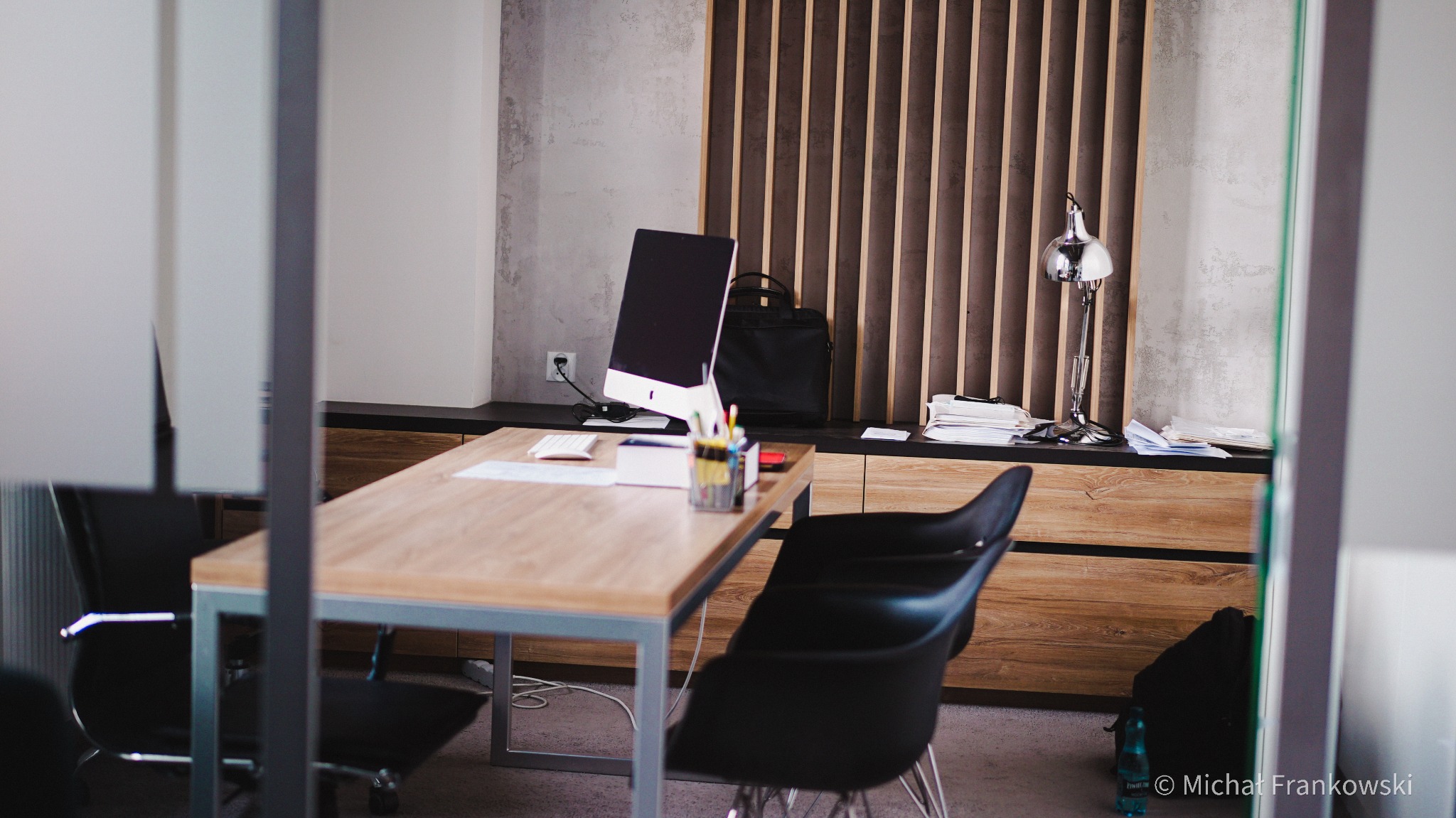 Nowoczesne biuro: drewniany stół z komputerem, lampką i przyborami biurowymi, czarne krzesła, widok przez szklaną ścianę.
