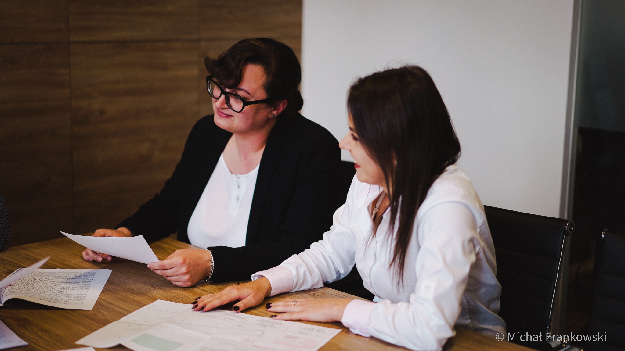 Dwie kobiety w biurze omawiają dokumenty na drewnianym stole, jedna trzyma arkusz papieru, druga opiera dłonie na planach.