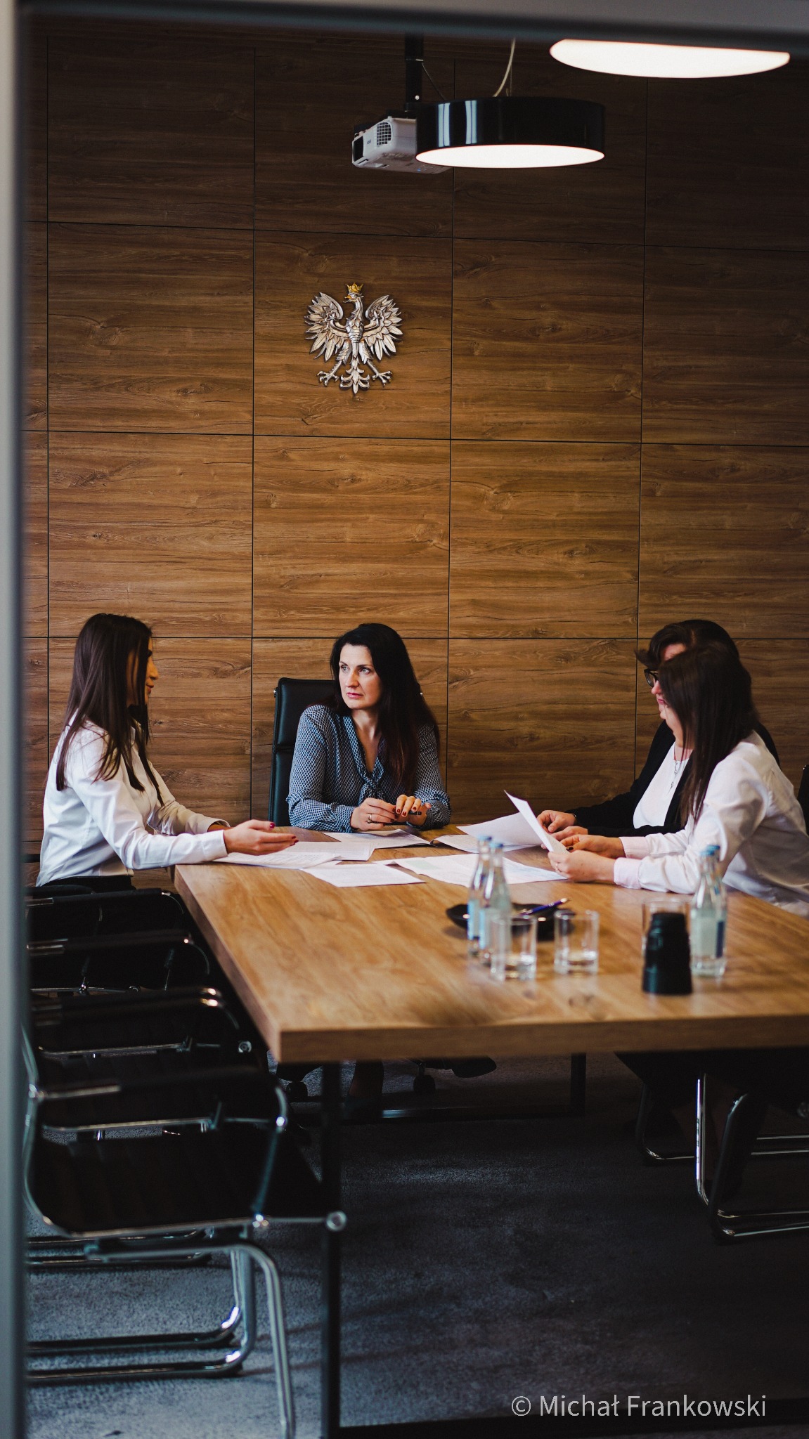 Spotkanie w kancelarii: cztery osoby przy drewnianym stole, dokumenty, woda w szklankach i butelkach, na ścianie godło Polski, oświetlenie sufitowe i projektor.