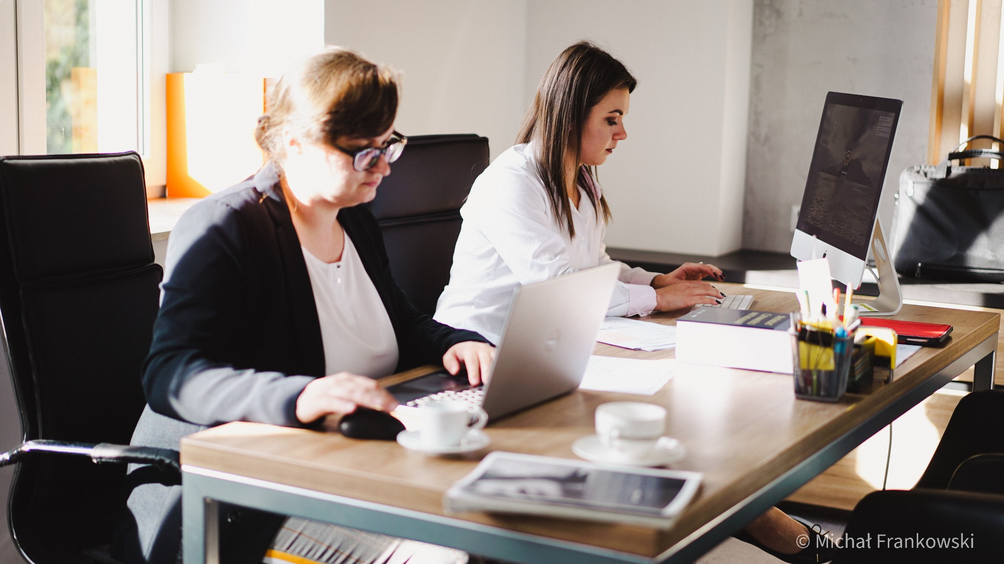Dwie kobiety pracują przy biurku w jasnym biurze, używając laptopa i komputera stacjonarnego, z filiżankami kawy na stole.