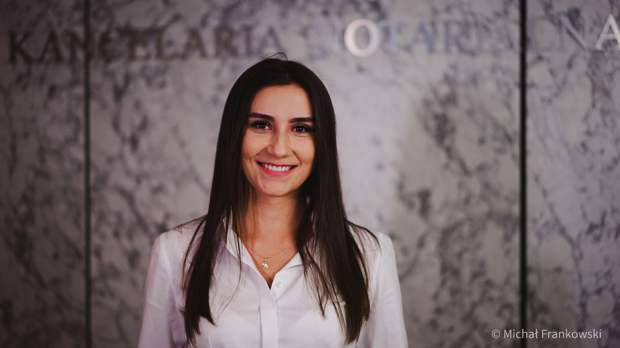 Uśmiechnięta kobieta z długimi, ciemnymi włosami w białej koszuli na tle marmurowej ściany z napisem 'Kancelaria Notarialna'.