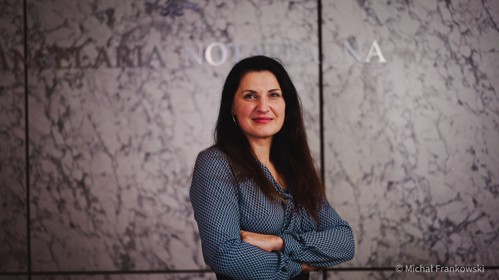 Portret kobiety z założonymi rękami na tle marmurowej ściany z napisem 'Kancelaria Notarialna'.