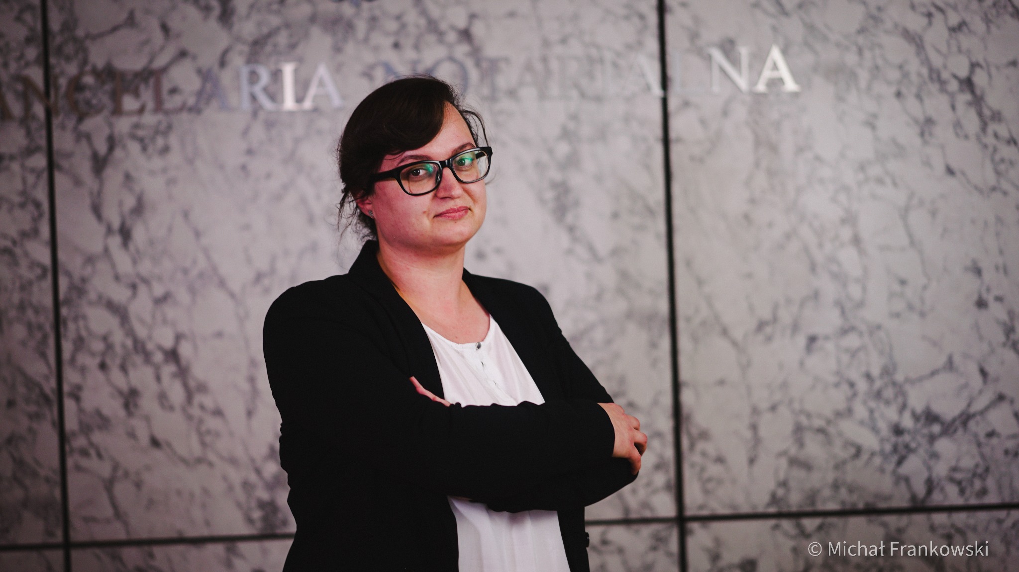 Kobieta w okularach i czarnej marynarce, z założonymi rękami na tle marmurowej ściany z napisem 'Kancelaria Notarialna'.