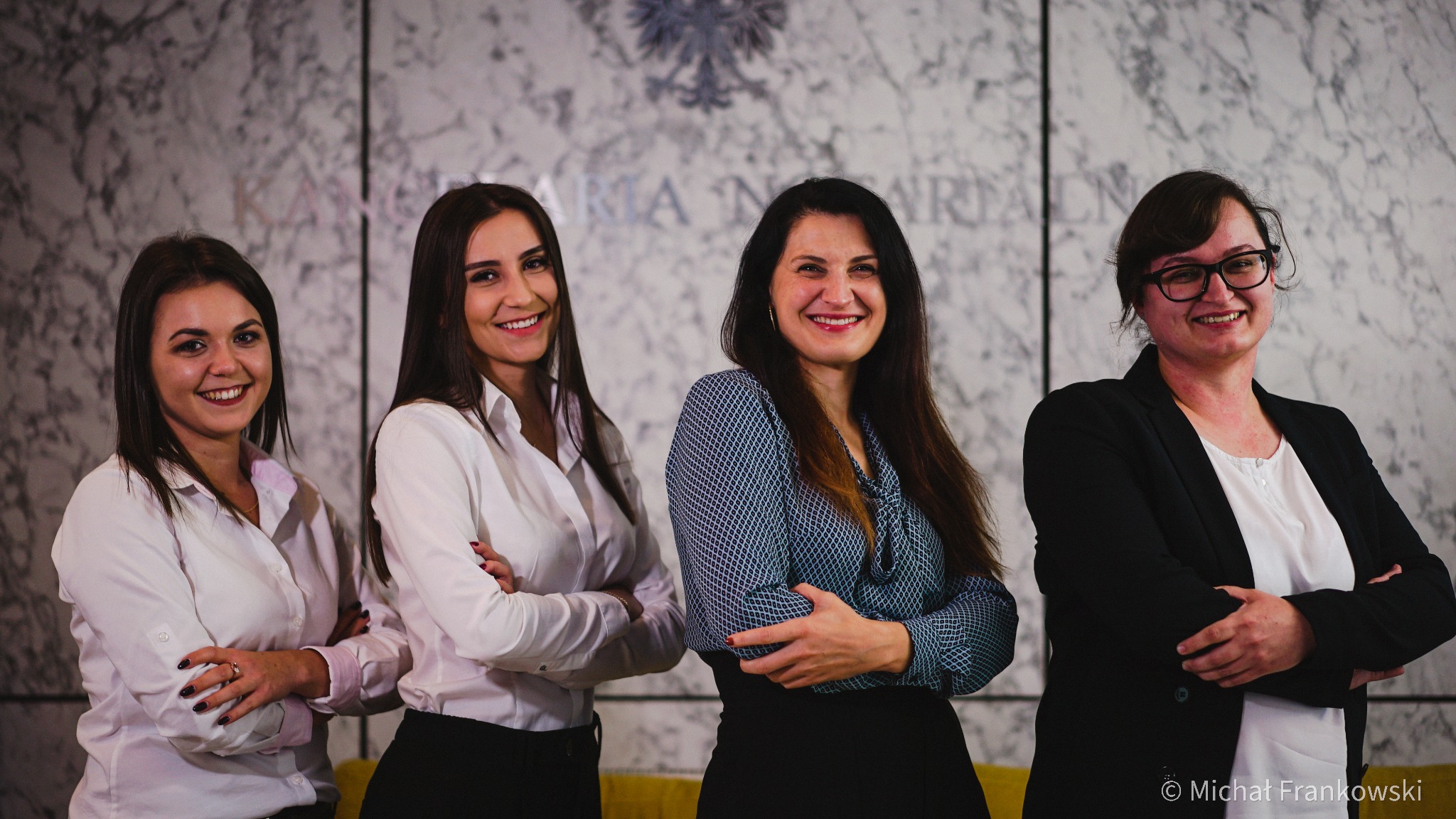 Zespół notarialny: cztery uśmiechnięte kobiety w eleganckich strojach stojące na tle marmurowej ściany z godłem państwowym i napisem 'Izba Notarialna'