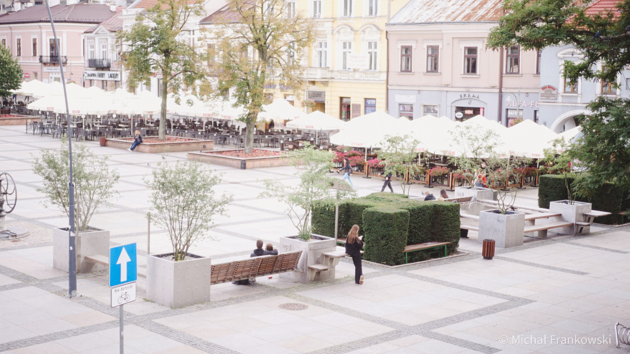Miejski plac z restauracjami pod parasolami, ławkami z donicami i strzyżonymi żywopłotami, widok z góry.