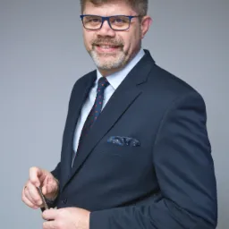 Uśmiechnięty mężczyzna w garniturze, okularach w niebieskich oprawkach i krawacie w kropki, trzymający w dłoni długopis na tle szarego tła.