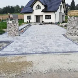 Ułożona szara kostka brukowa przed domem z białymi ścianami, z obramowaniem z ciemniejszej kostki i gabionami po bokach, widoczne przygotowane podłoże.