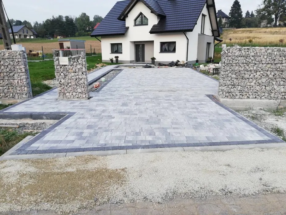 Ułożona szara kostka brukowa przed domem z białymi ścianami, z obramowaniem z ciemniejszej kostki i gabionami po bokach, widoczne przygotowane podłoże.