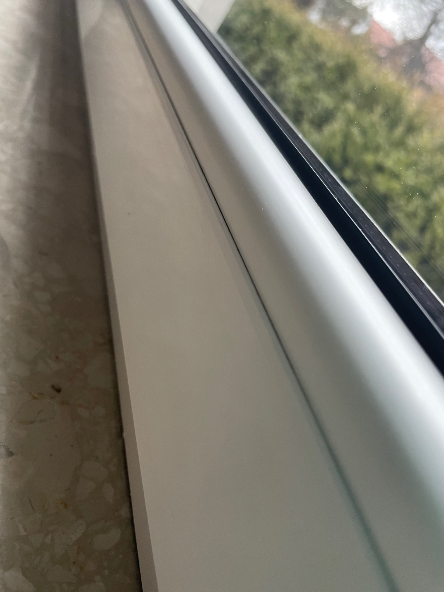 Fragment białego okna PCV z widokiem na zieleń ogrodu, uchwycony pod kątem, z widoczną fakturą parapetu.