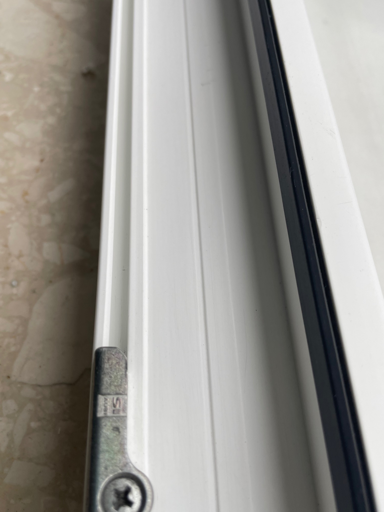 Fragment białej ramy okiennej z widocznym metalowym okuciem i śrubą na tle wzorzystego parapetu.