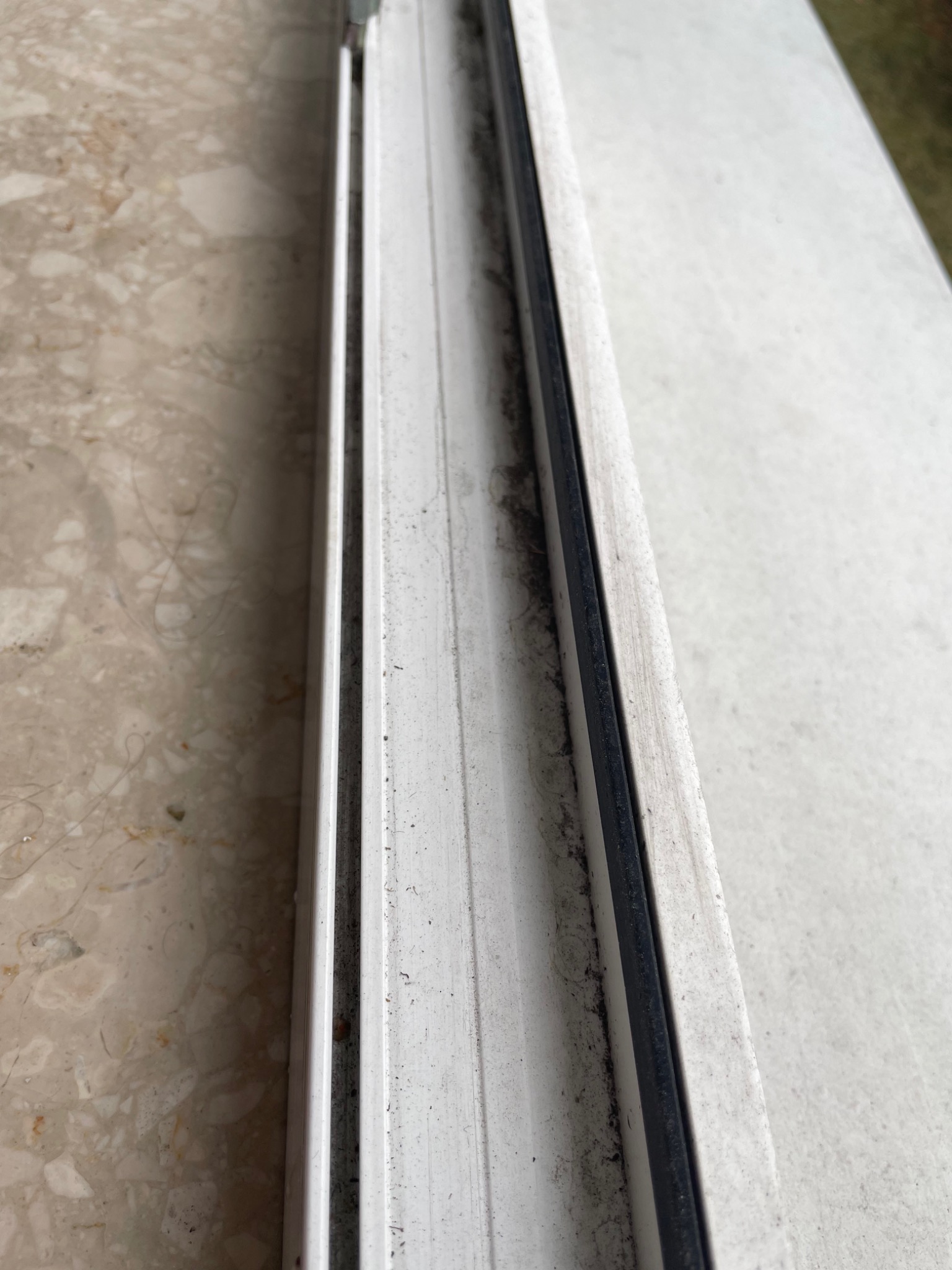 Detail zabrudzonego, białego profilu okiennego z czarną uszczelką, widoczne ślady kurzu i pleśni, marmurkowy parapet w tle.