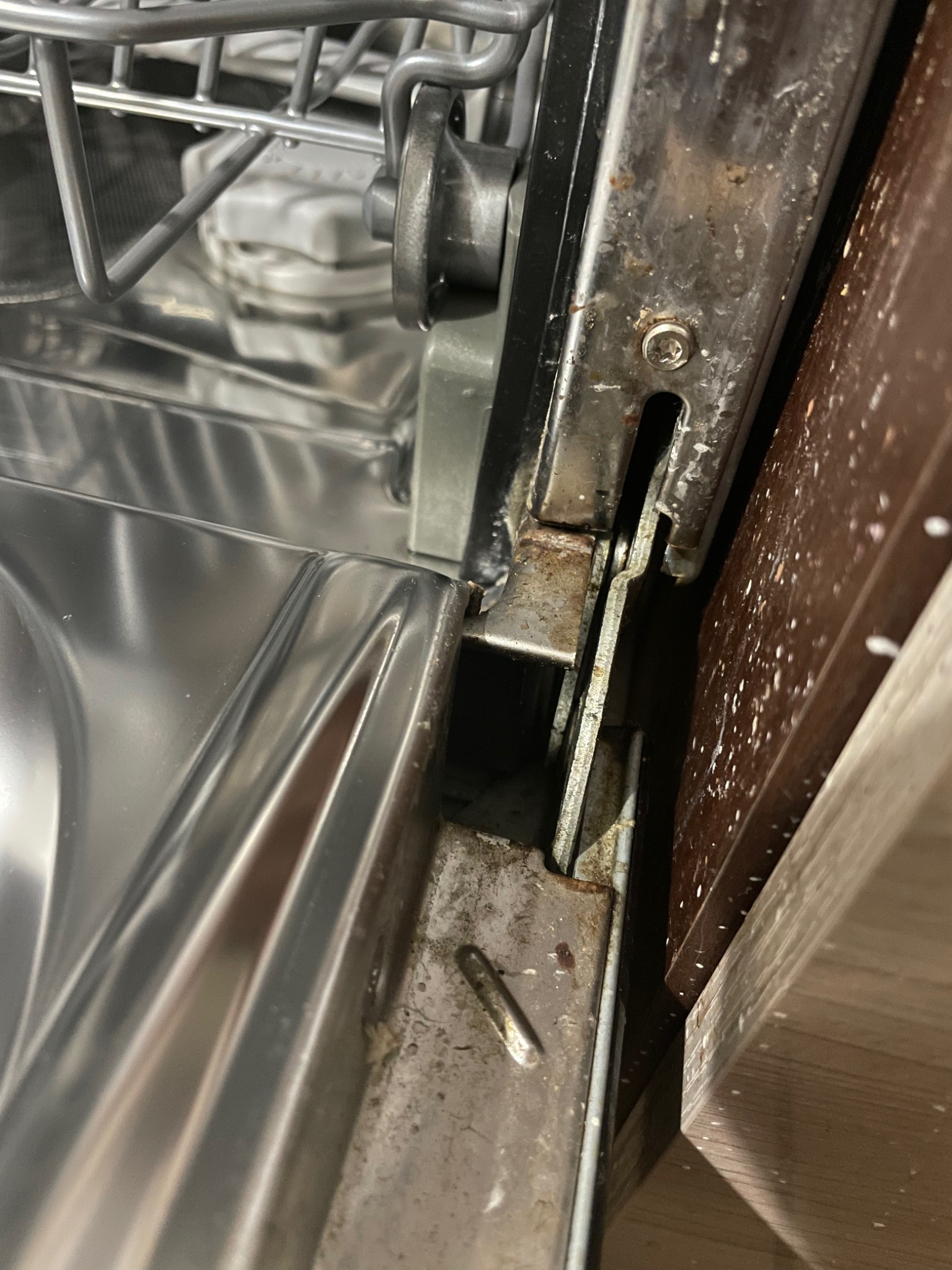 Szczegółowe ujęcie skorodowanych elementów mocujących drzwi zmywarki, widoczne ślady rdzy i zabrudzeń na metalowych częściach oraz obudowie zmywarki.
