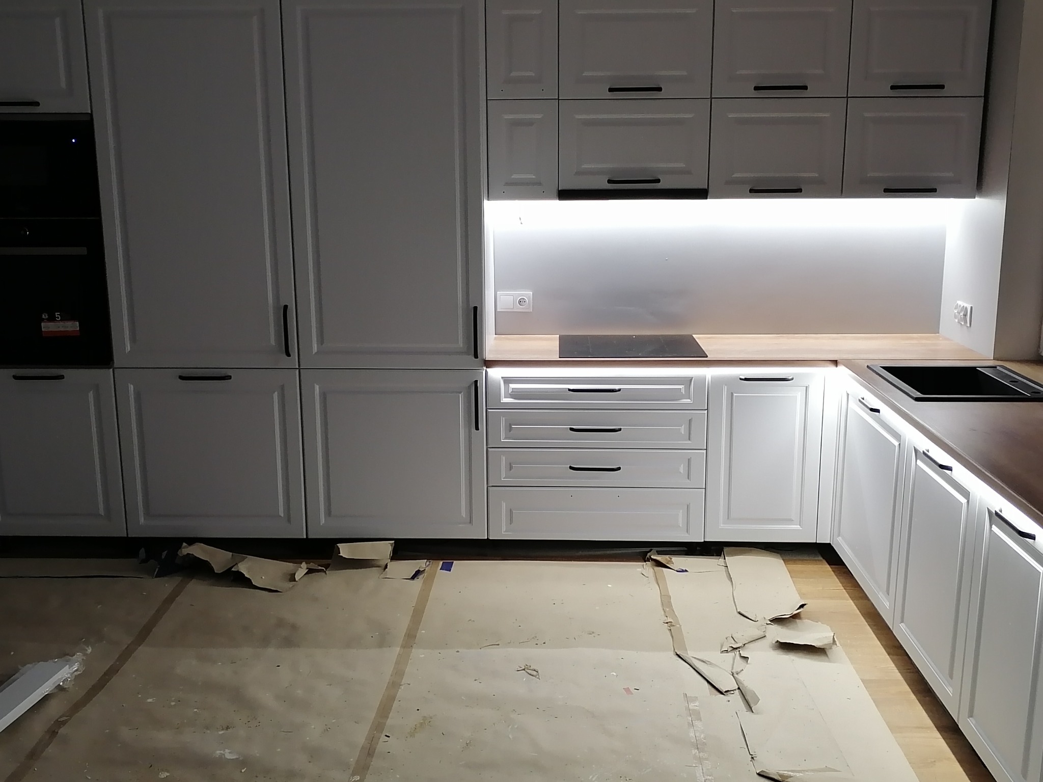 Biała zabudowa kuchenna z drewnianym blatem i czarnymi uchwytami, zabezpieczona kartonem na podłodze podczas montażu.