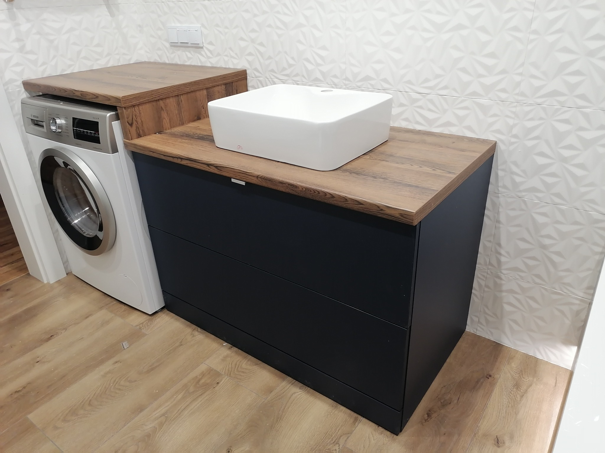 Nowoczesna łazienka z szafką pod umywalkę w kolorze antracytowym, blatem drewnopodobnym i białą, kwadratową umywalką nablatową, obok pralka.