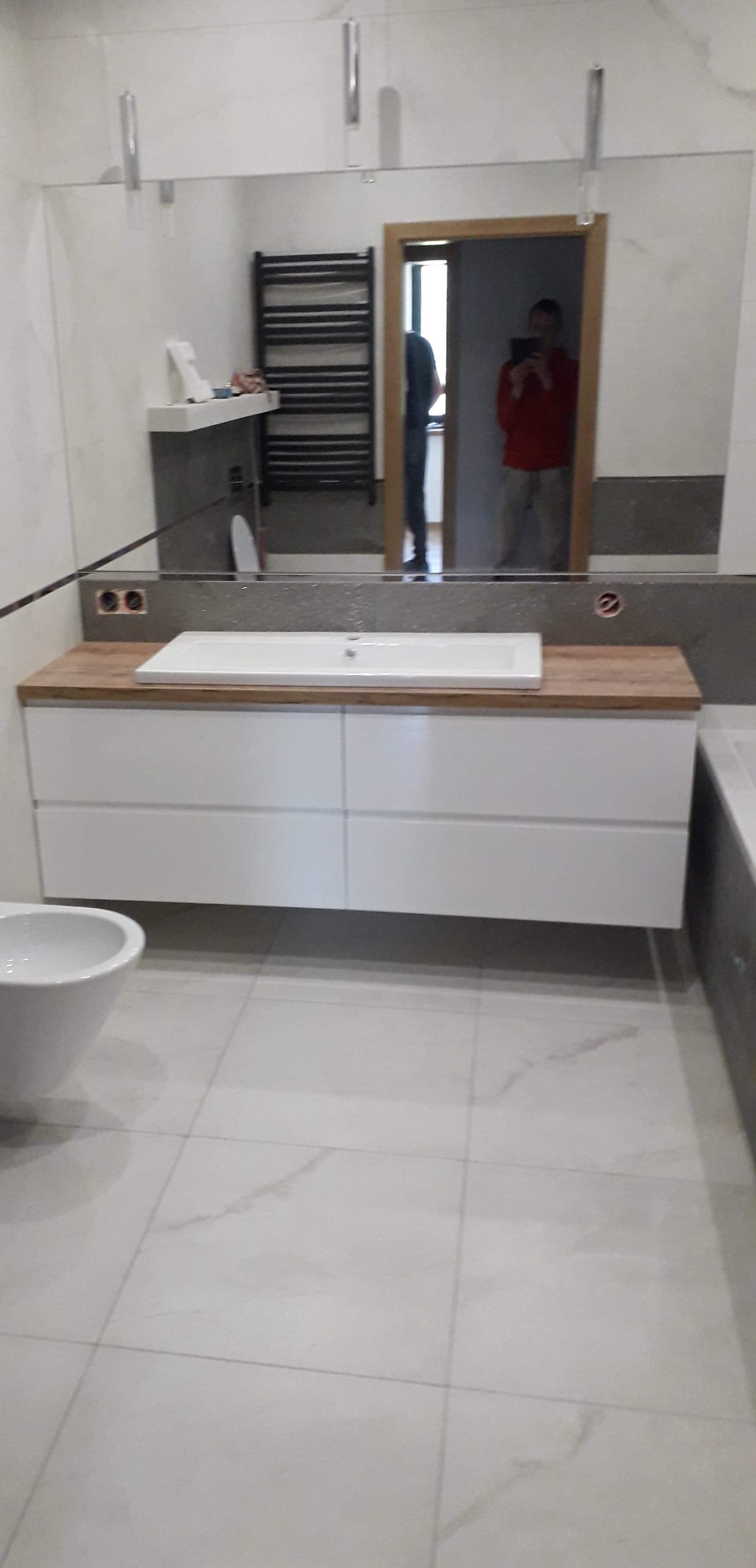 Nowoczesna łazienka z białą szafką pod umywalką z drewnianym blatem, duże lustro z oświetleniem, widoczne gniazdka elektryczne, bidet, oraz odbicie osoby robiącej zdjęcie w lustrze.