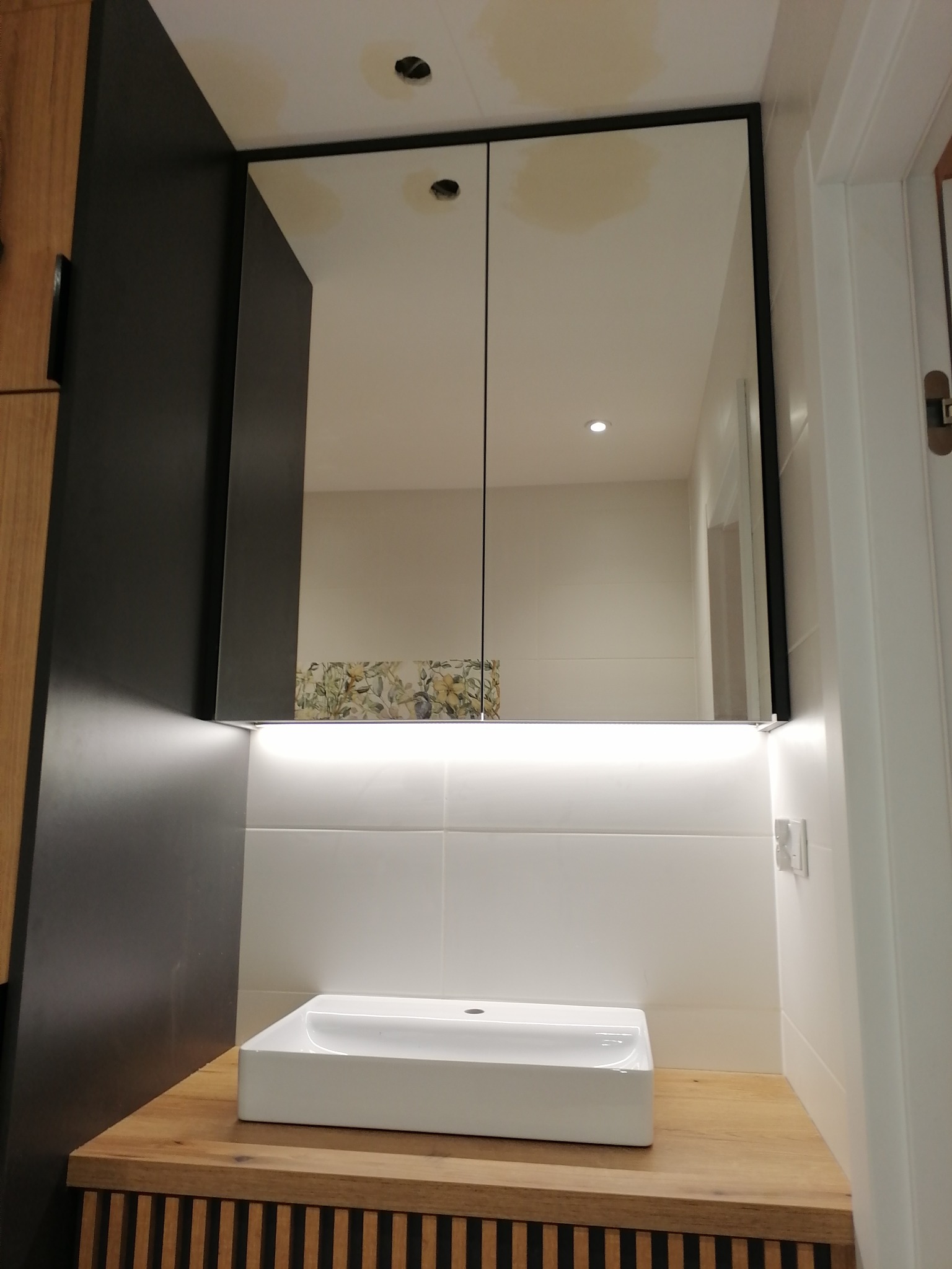 Nowoczesna łazienka z umywalką nablatową, szafką z drewnianym blatem i lustrem w czarnej ramie z podświetleniem LED, odbijającym fragment dekoracyjnej tapety.