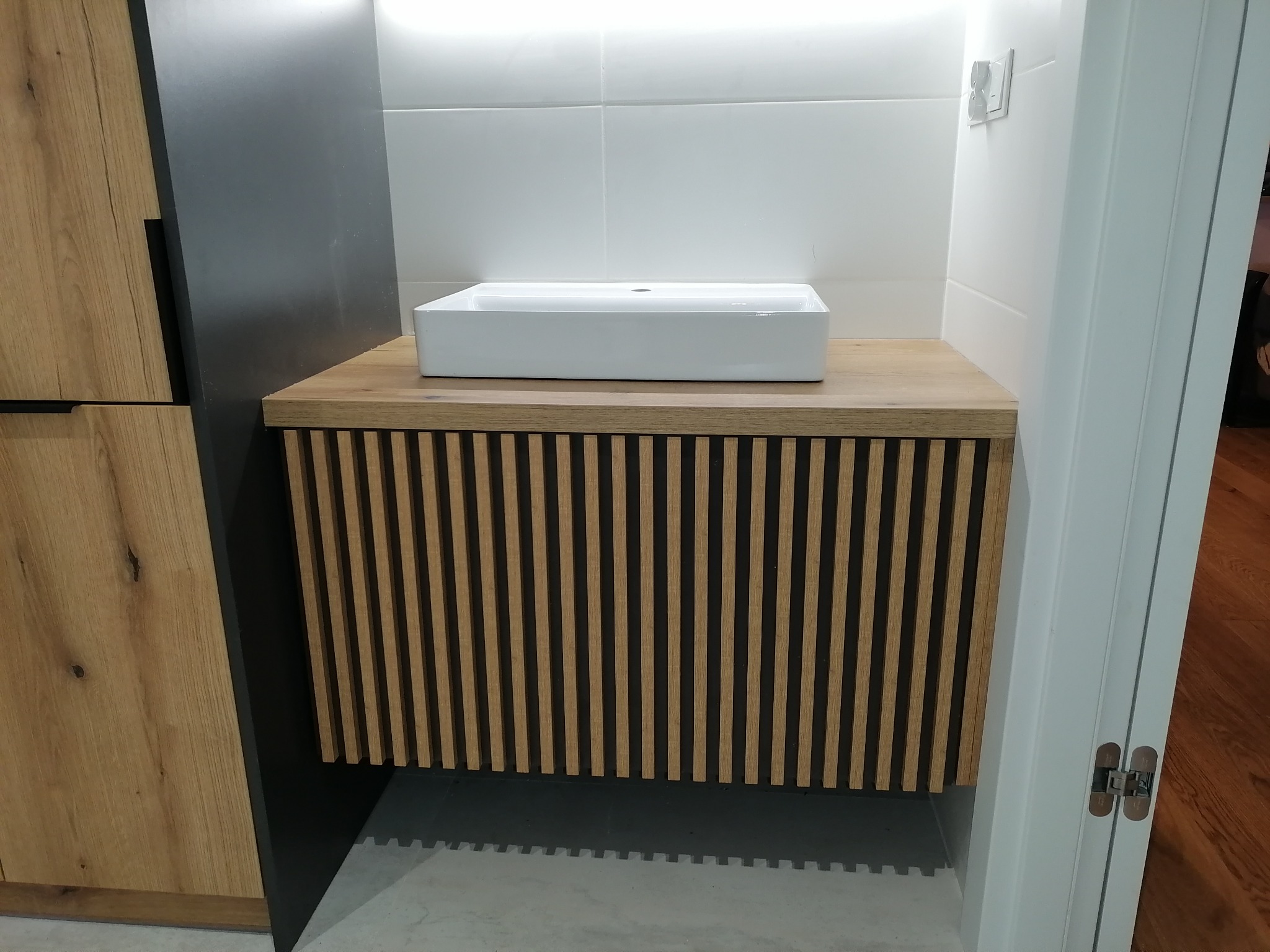 Nowoczesna łazienka z prostokątną umywalką nablatową, szafką z drewnianymi lamelami i fragmentem drewnianej szafy po lewej stronie.