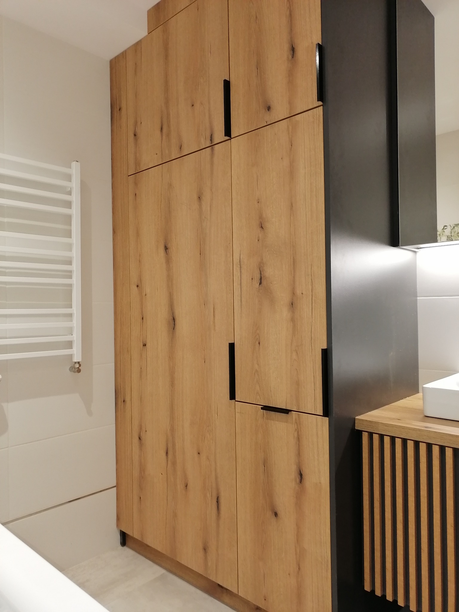 Wysoka szafa z frontami w odcieniach drewna i czarnymi uchwytami w nowoczesnej łazience, obok grzejnik drabinkowy i umywalka z blatem.