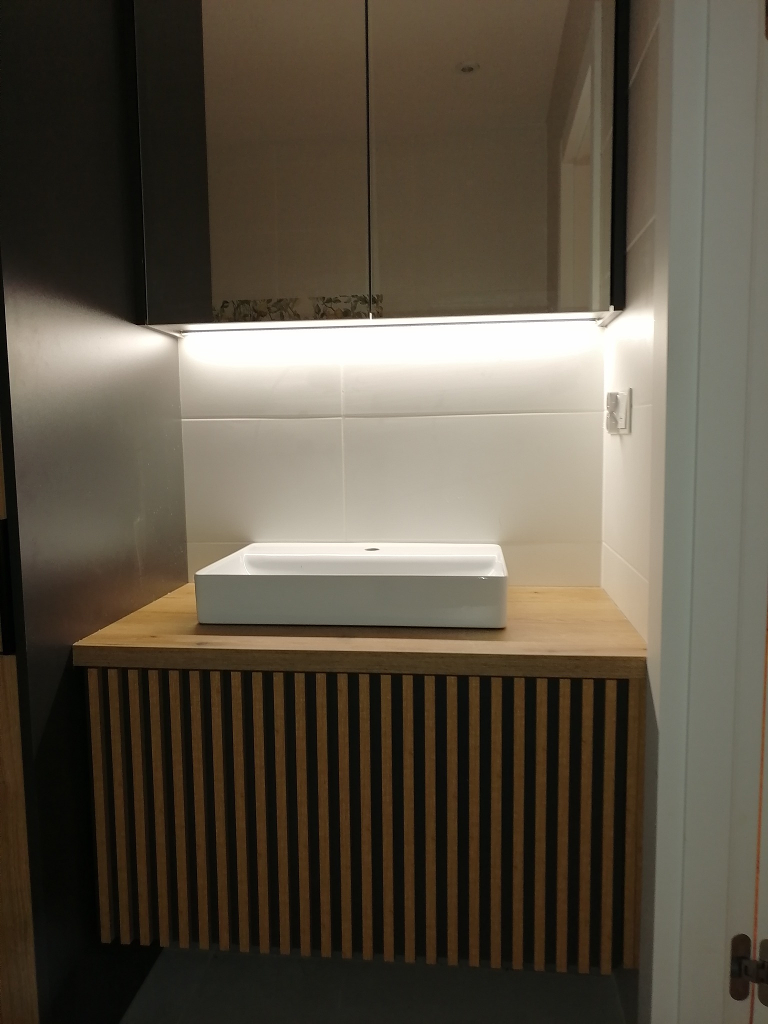 Nowoczesna umywalka nablatowa o kwadratowym kształcie na szafce z frontem z pionowych listewek, oświetlona od góry listwą LED, widok z góry na lustrzane szafki.