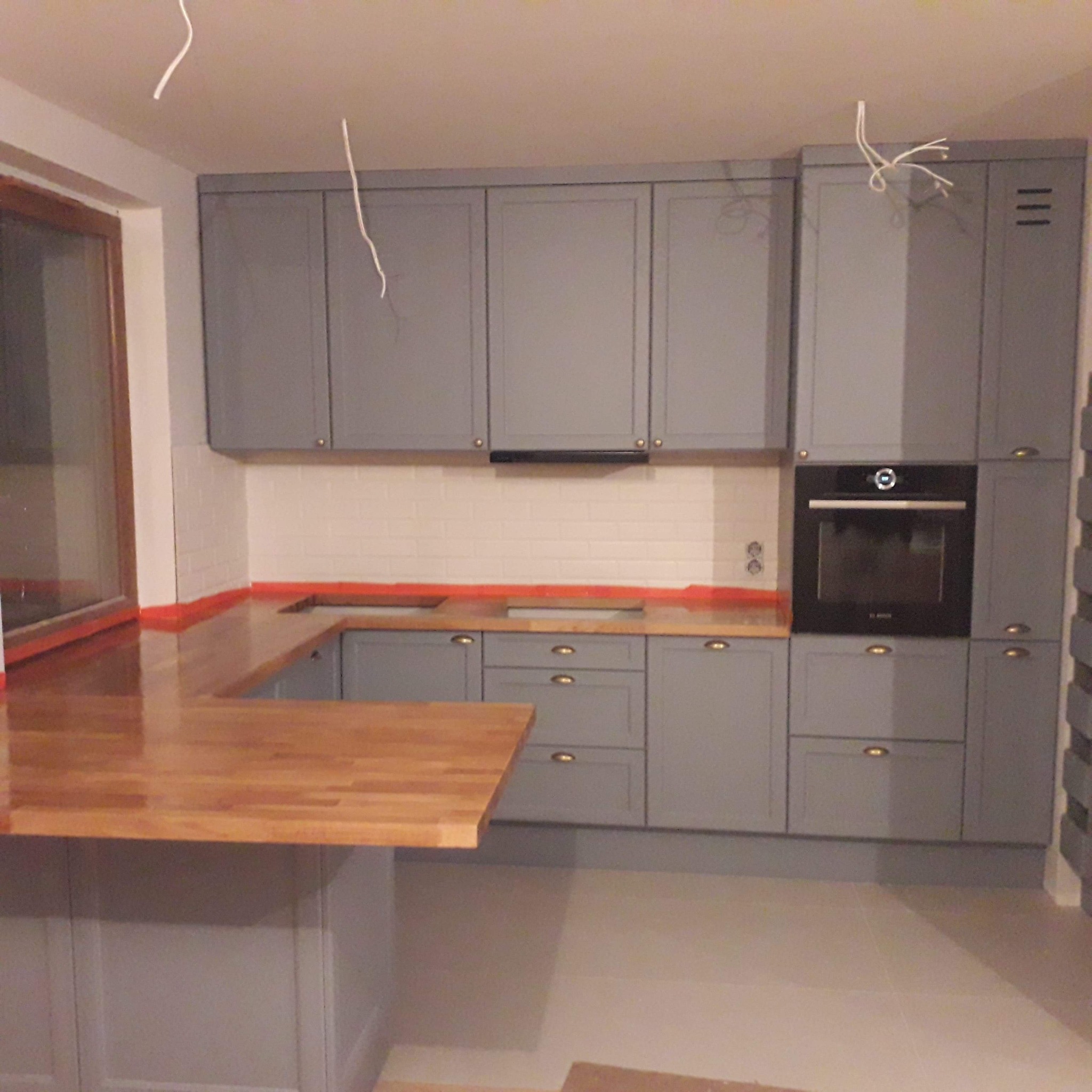 Szara zabudowa kuchenna z drewnianym blatem i wyspą, widoczne kable zwisające z sufitu, białe płytki na ścianie i zabudowana piekarnik