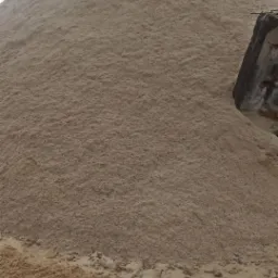 Góra jasnobrązowego materiału sypkiego, prawdopodobnie agropellet lub nawóz, z fragmentem betonowej konstrukcji w tle, sfotografowana w pochmurny dzień.