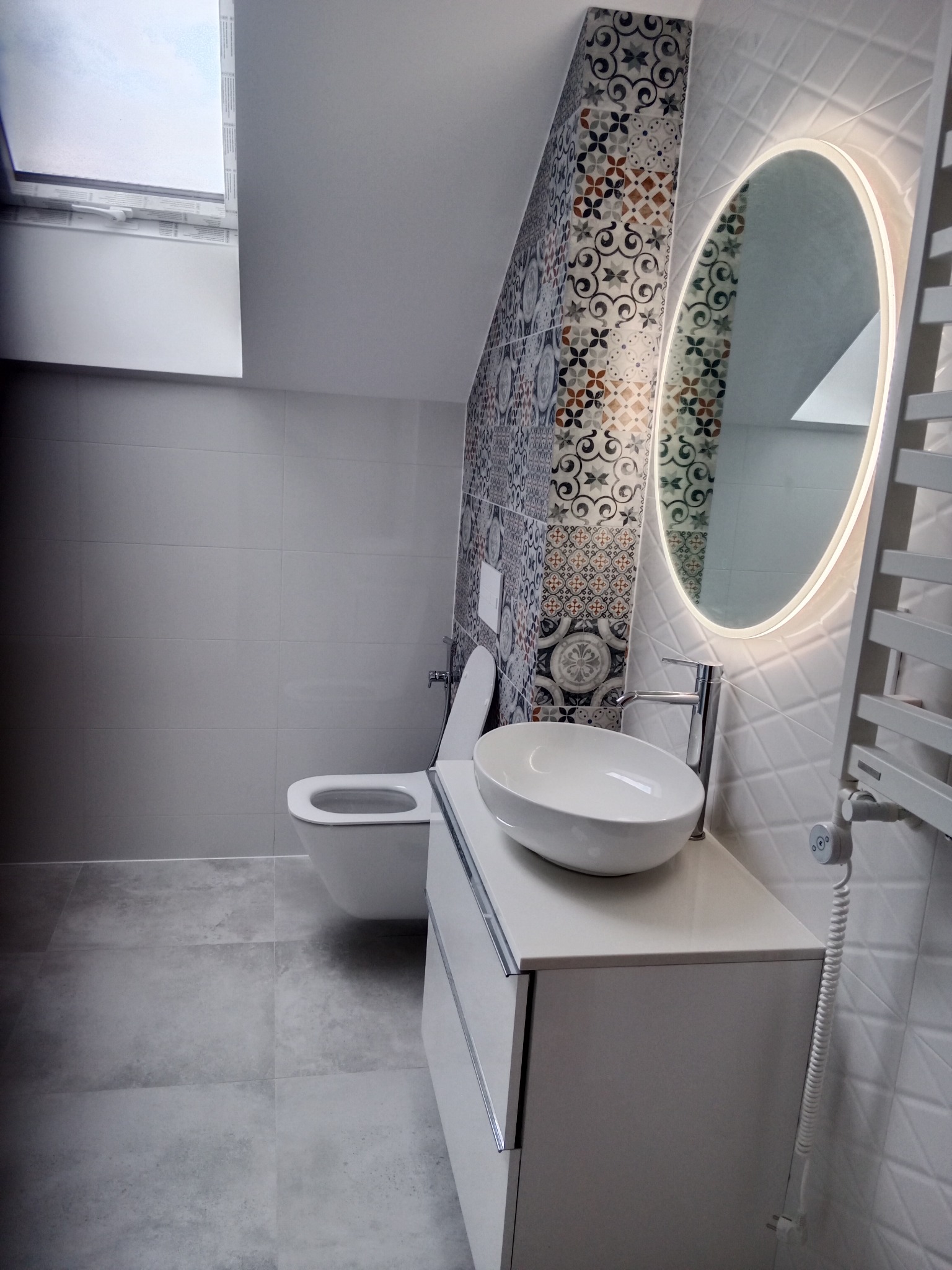 Nowoczesna łazienka na poddaszu z oknem dachowym, szarą płytką podłogową, wiszącą toaletą, umywalką nablatową na białej szafce i podświetlanym lustrem owalnym oraz dekoracyjną ścianą...