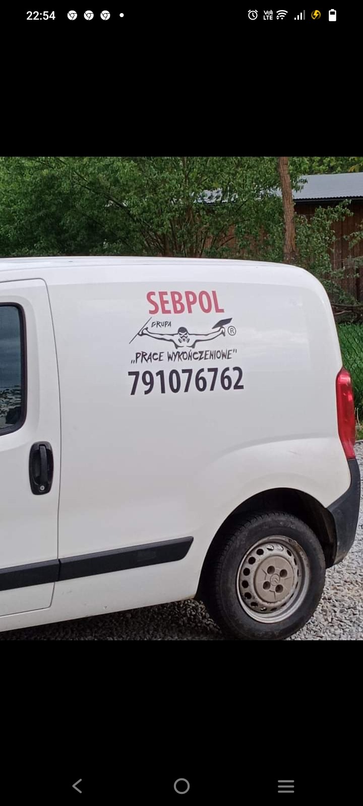 Biały samochód dostawczy z logo firmy SEBPOL GRUPA 'PRACE WYKOŃCZENIOWE' i numerem telefonu 791076762 na boku, zaparkowany na żwirowym podłożu, w tle zieleń drzew.
