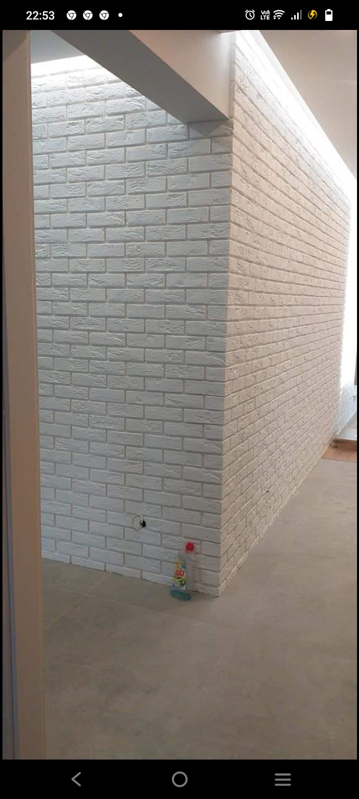 Ściana wykończona białą cegłą dekoracyjną w nowo wyremontowanym pomieszczeniu, widoczny fragment podłogi z płytek i butelka płynu do czyszczenia w rogu.