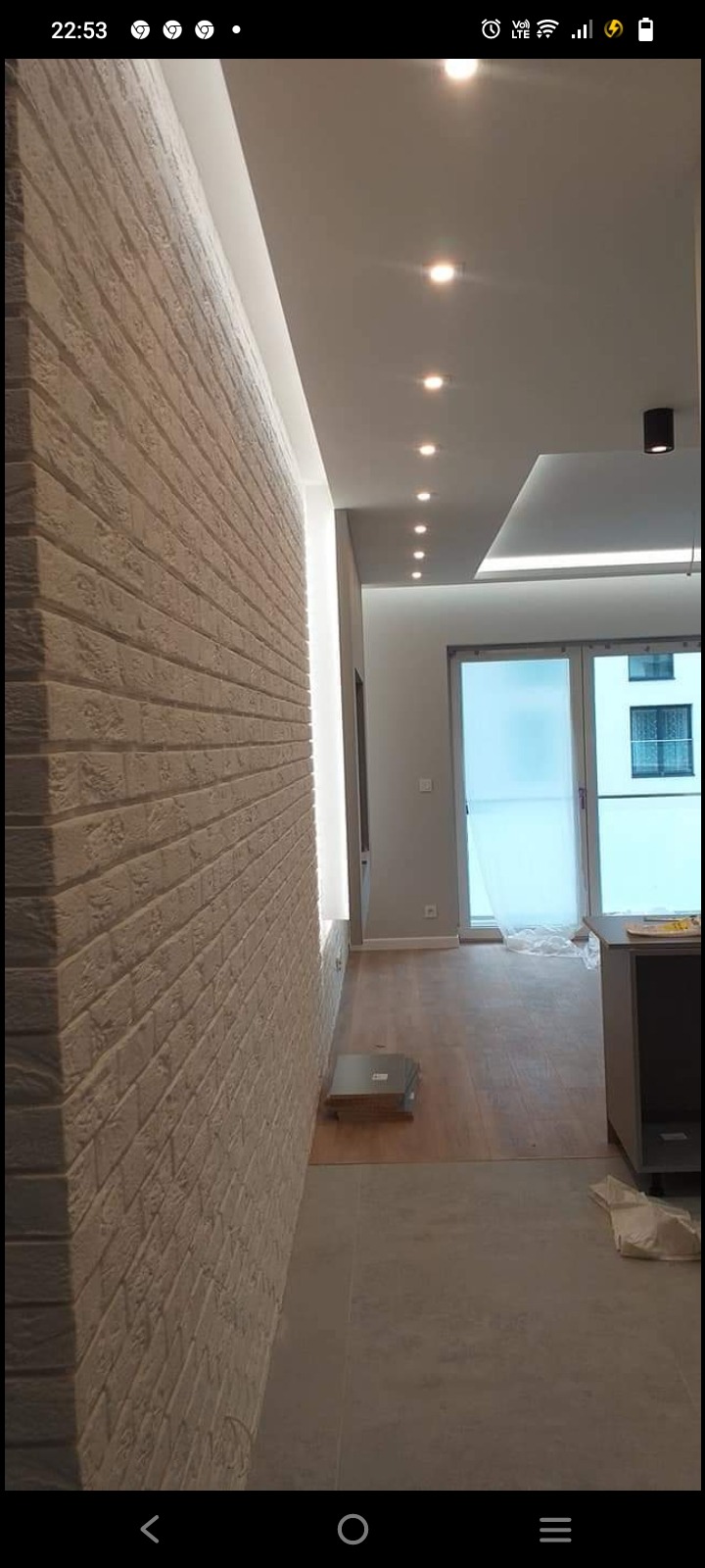 Wykończona ściana z cegły dekoracyjnej w jasnym odcieniu, oświetlona listwą LED, z widocznym fragmentem sufitu z punktowym oświetleniem i podłogą wyłożoną panelami oraz płytkami w nowocześnie...