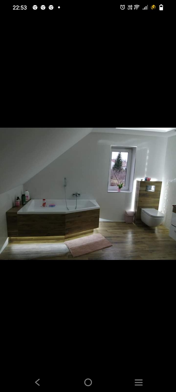 Ukończona łazienka na poddaszu z wanną narożną obudowaną drewnem, podświetlanym ledowo, drewnianą podłogą, wiszącą toaletą i oknem z widokiem na drzewo.