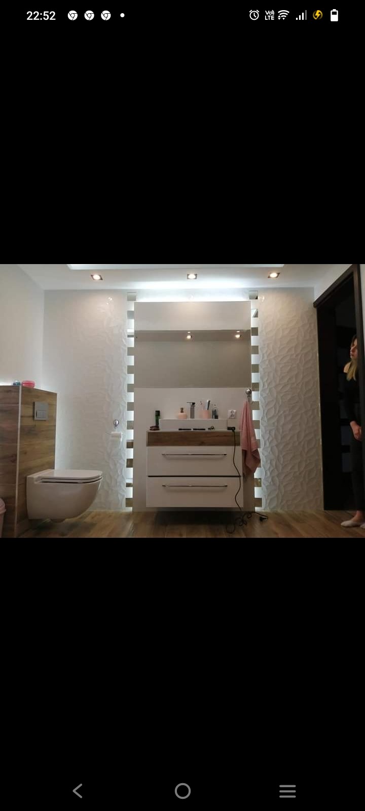 Nowoczesna łazienka z podwieszaną toaletą, szafką z umywalką i lustrem podświetlanym listwami LED, wykończona płytkami z trójwymiarową fakturą i drewnopodobną podłogą.