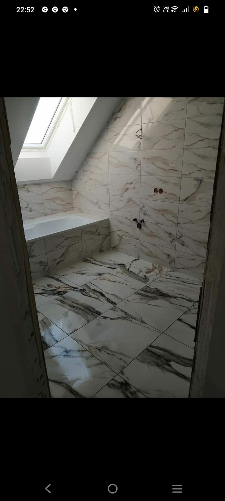 Wykończona łazienka na poddaszu z marmurowymi płytkami, widoczna wanna pod oknem dachowym i instalacja wodna bez armatury.