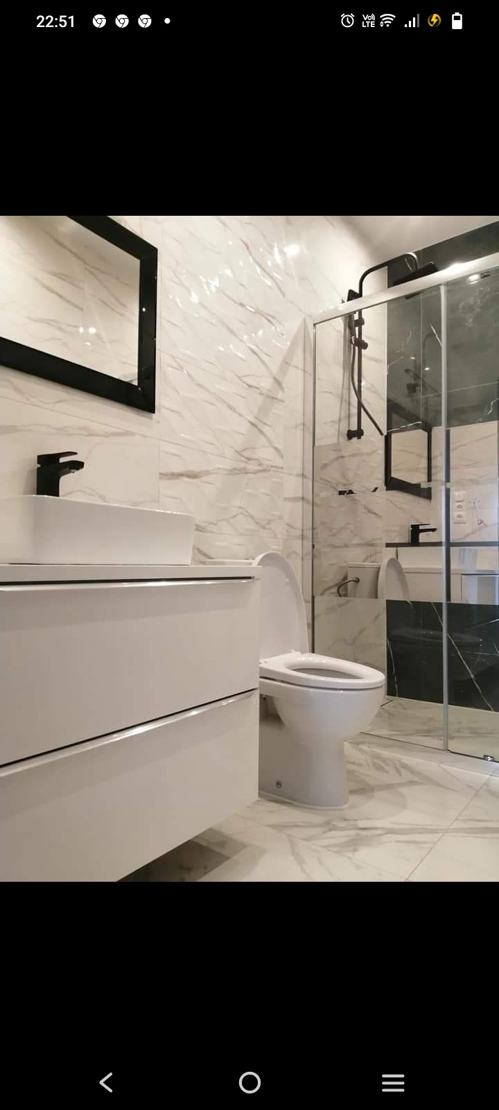 Nowoczesna łazienka z białymi marmurkowymi płytkami, czarną armaturą, wiszącą toaletą i kabiną prysznicową typu walk-in z lustrem.