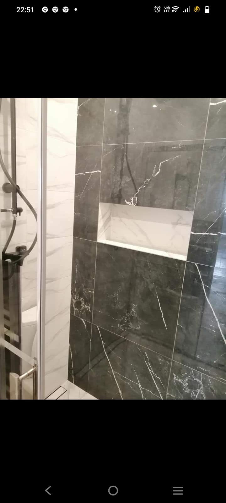 Nowoczesna kabina prysznicowa z czarnym, marmurkowym wykończeniem ścian i wnęką na kosmetyki, widok z boku na szklane drzwi i czarną armaturę.