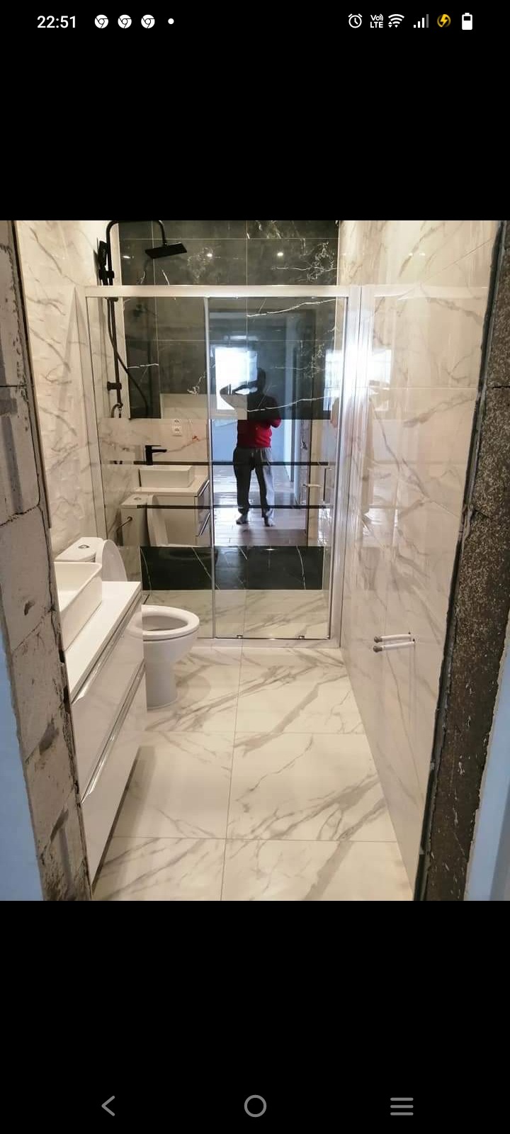 Nowoczesna łazienka z kabiną prysznicową ze szklanymi przesuwnymi drzwiami, białą umywalką z szafką i toaletą, wykończona białymi płytkami imitującymi marmur z czarnymi akcentami.