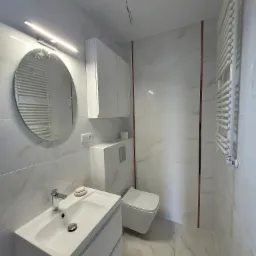 Nowoczesna łazienka w jasnych barwach z marmurkowymi płytkami, okrągłe lustro z podświetleniem LED, biała umywalka z szafką, wisząca toaleta i grzejnik drabinkowy.