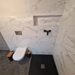 Nowoczesna łazienka po remoncie: białe płytki imitujące marmur, czarny brodzik bezprogowy z odpływem liniowym, podwieszana toaleta z drewnianym pojemnikiem na szczotkę, wnęka w ścianie wykończona...