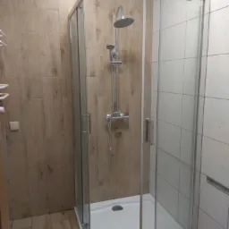 Nowoczesna kabina prysznicowa z chromowaną armaturą na białym brodziku, otoczona płytkami imitującymi drewno po jednej stronie i białymi płytkami po drugiej, z niebieską taśmą malarską...