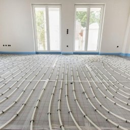 Aqua team Dawid Foerster - Białe rury ogrzewania podłogowego ułożone spiralnie na szarej macie izolacyjnej w jasnym pomieszczeniu z dwoma oknami i zabezpieczoną folią ścianą.