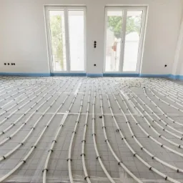 Białe rury ogrzewania podłogowego ułożone spiralnie na szarej macie izolacyjnej w jasnym pomieszczeniu z dwoma oknami i zabezpieczoną folią ścianą.