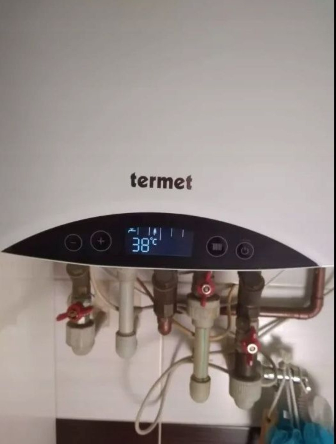Biały piec gazowy marki Termet z wyświetlaczem wskazującym temperaturę 38 stopni Celsjusza, widoczne rury i zawory instalacji grzewczej na tle płytek ceramicznych.
