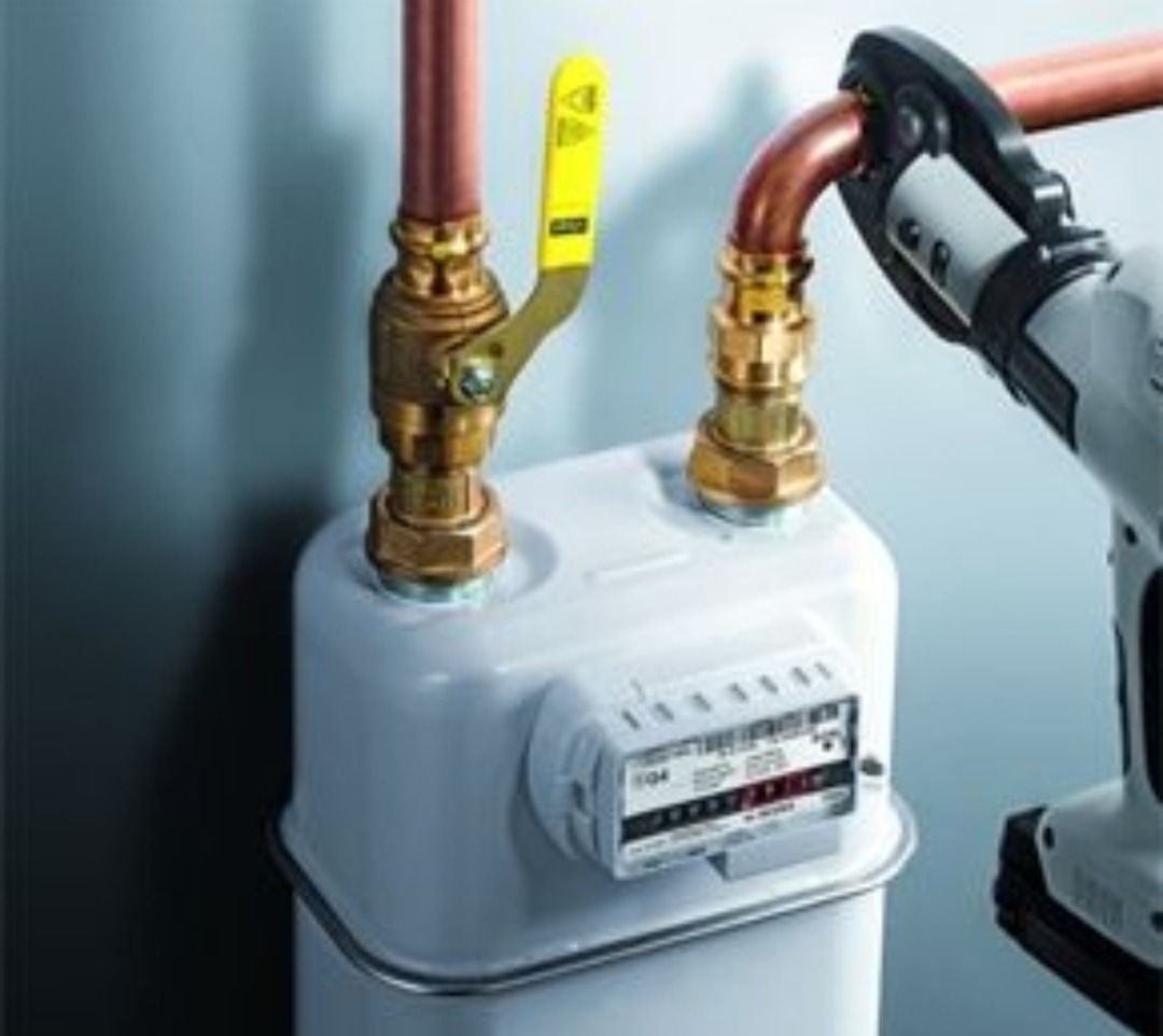 Podłączenie miedzianej rury gazowej do białego gazomierza za pomocą zaciskarki hydraulicznej, widoczny zawór odcinający z żółtą rączką.