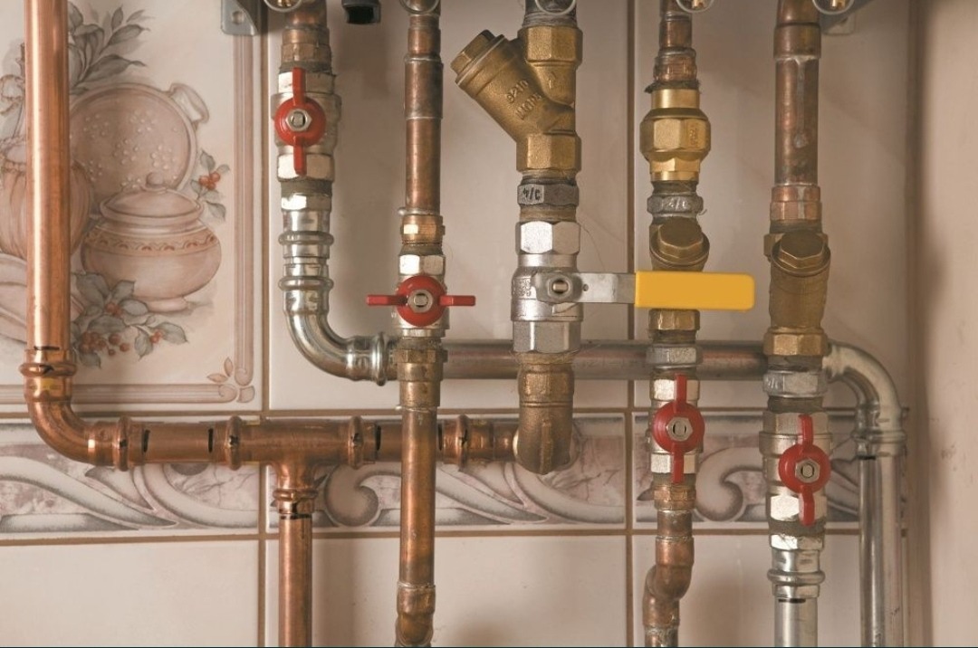 Złożony system rur miedzianych i mosiężnych z zaworami kulowymi z czerwonymi pokrętłami, filtrem siatkowym i zaworem z żółtą dźwignią, zamontowany na tle płytek ceramicznych z motywem kuchennym.