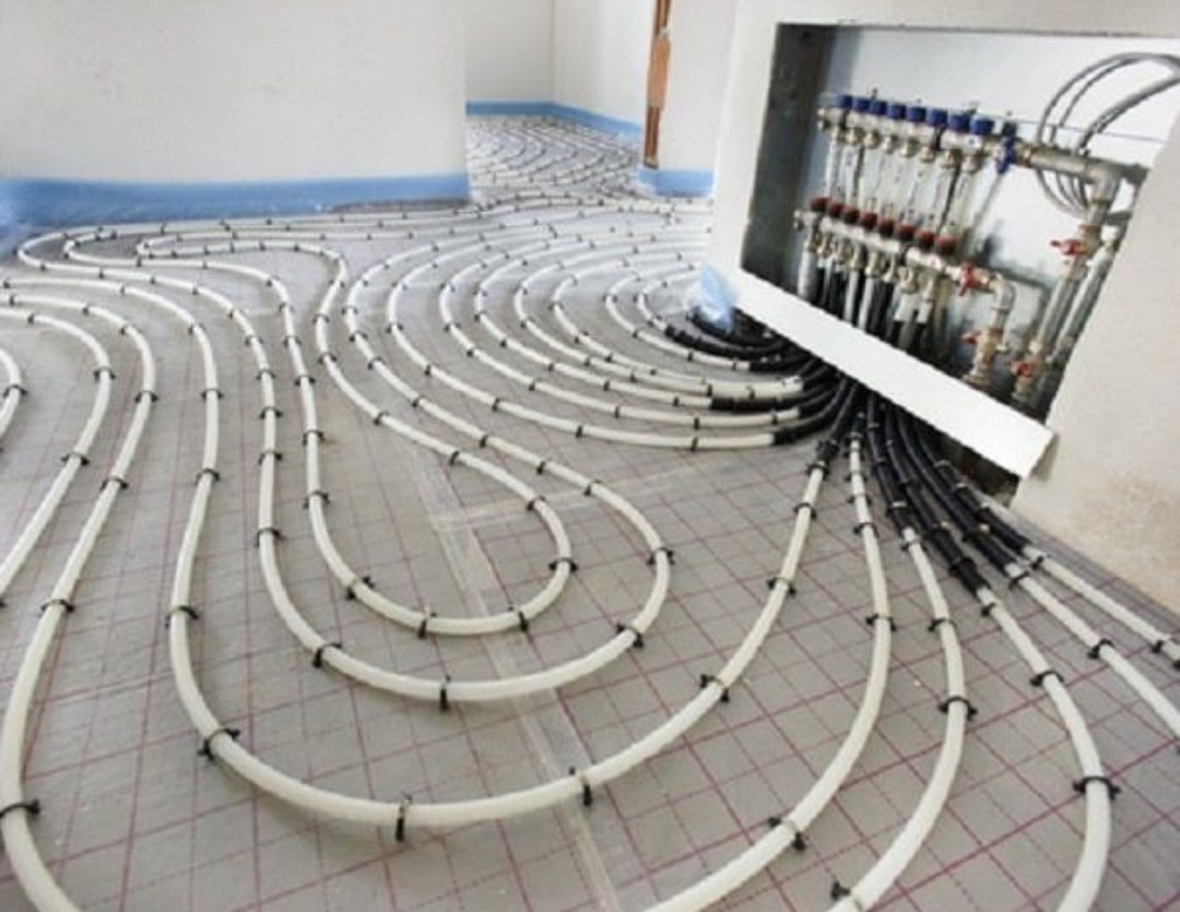 Rozłożone białe rury ogrzewania podłogowego na siatce, widoczny rozdzielacz z zaworami w ścianie.